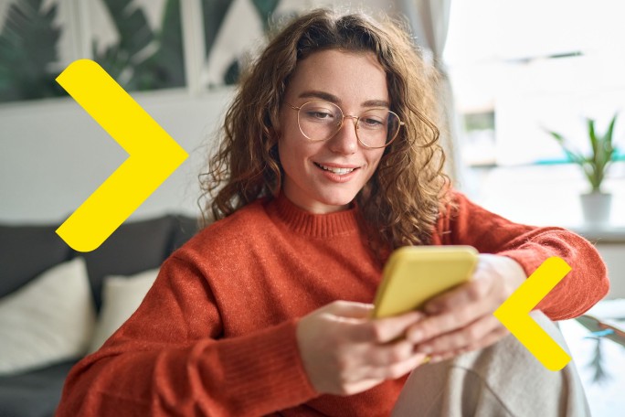 Credit de nevoi personale. O persoana sta pe o canapea, purtand un pulover portocaliu si tinand un smartphone galben. In fundal se afla o planta si tablouri inramate pe perete.