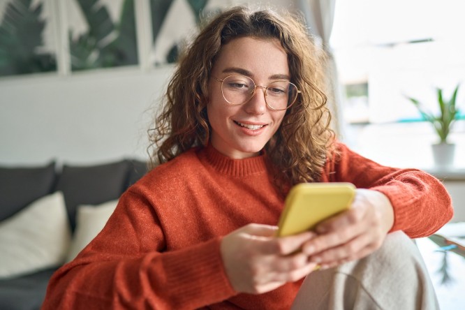 O persoana sta pe o canapea, purtand un pulover portocaliu si tinand un smartphone galben. In fundal se afla o planta si tablouri inramate pe perete.