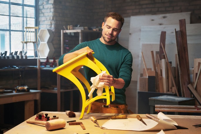 Un meșter este surprins în timp ce lustruiește un scaun galben intens într-un atelier de tâmplărie. Decorul include unelte, scânduri de lemn și schițe de design pe masă, evidențiind un spațiu creativ. Peretele din cărămidă și fereastra mare oferă o atmosferă rustică și bine luminată. Accentul este pus pe măiestria și atenția la detalii în realizarea mobilierului.