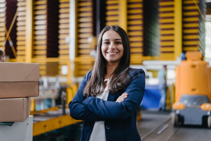 O femeie stă încrezătoare cu brațele încrucișate într-un depozit industrial modern. În fundal se observă cutii stivuite, utilaje galbene și echipamente automatizate. Scena evidențiază un mediu profesional cu elemente vizuale avansate de logistică și producție.