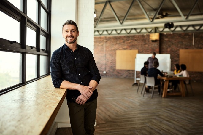 O persoana se sprijina de pervazul unei ferestre intr-o camera cu ferestre mari, pereti din caramida expusa si tavan in stil industrial. In fundal, un grup de persoane sta in jurul unei mese, iar o persoana sta in picioare langa o tabla alba.